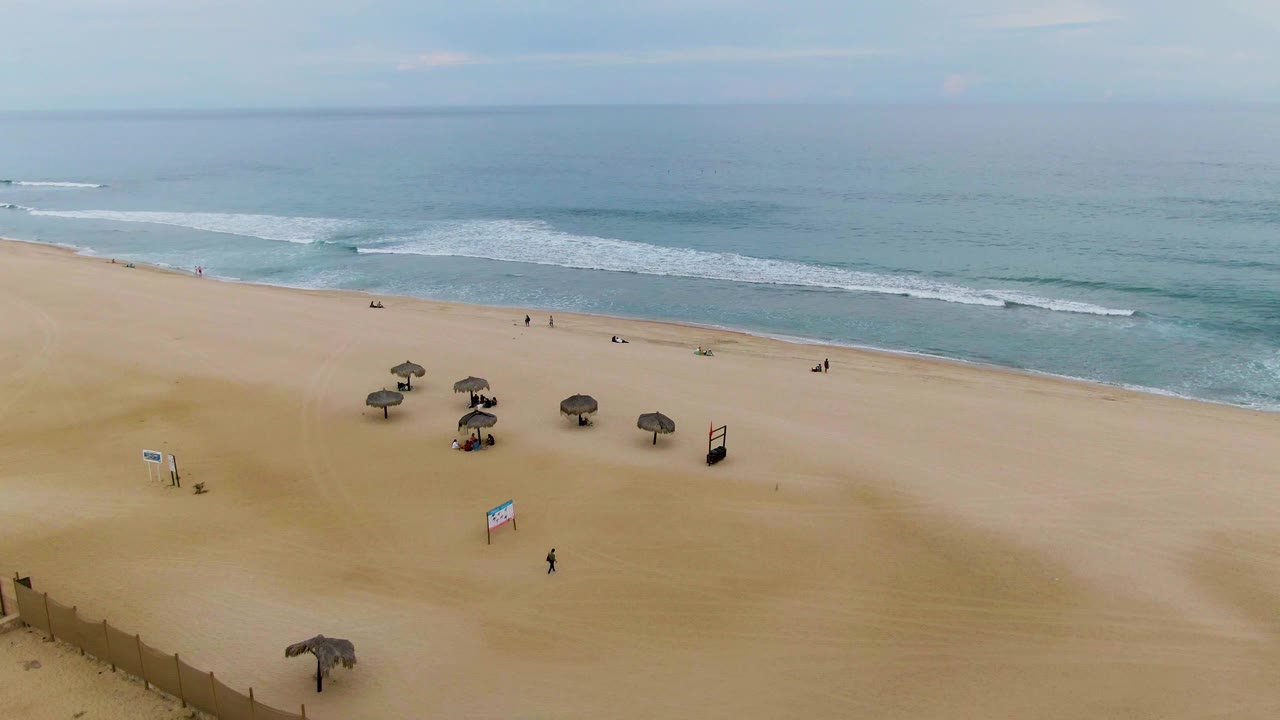 Sandy beach with thatched umbrellas, scattered people, surf, and horizon.