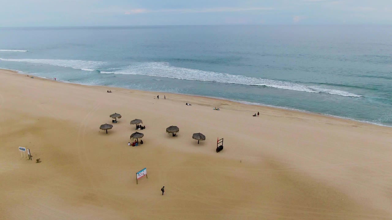 Wide aerial view of the San Jose Del Cabo shoreline with fewer fence elements visible, thatched umbrellas on the sand, distant people, small signs, and layered white waves offshore.