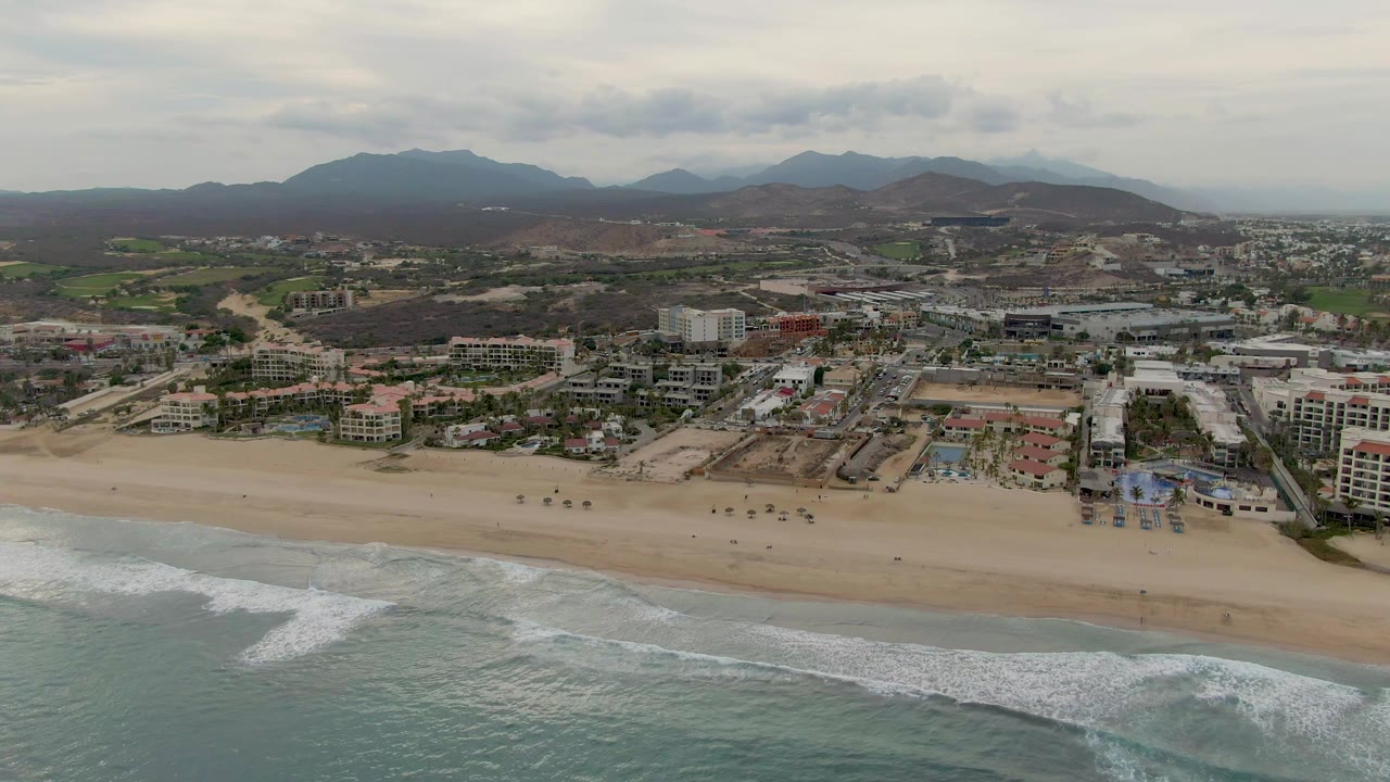 San Jose del Cabo aerial view with white wave foam spread across the lower shore, open sand in the center, clustered coastal buildings inland, hazy mountains, and no visible people.