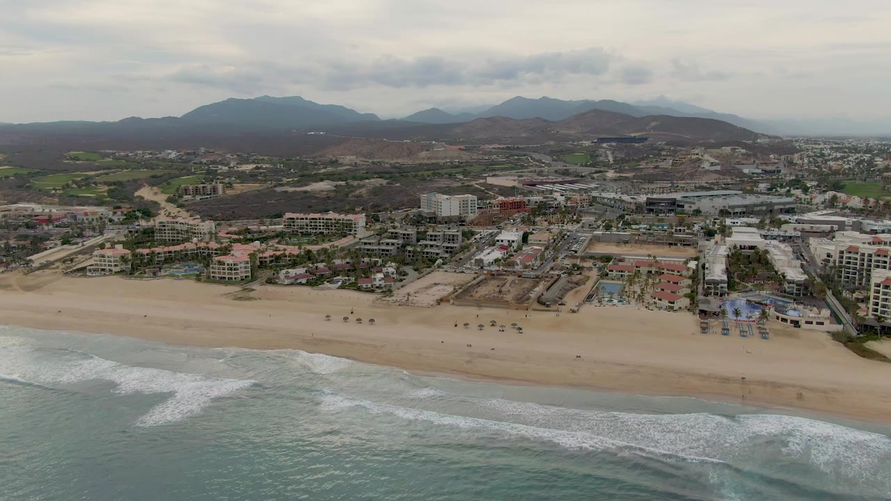 Aerial view of surf along a broad beach with coastal buildings and dark hills behind.