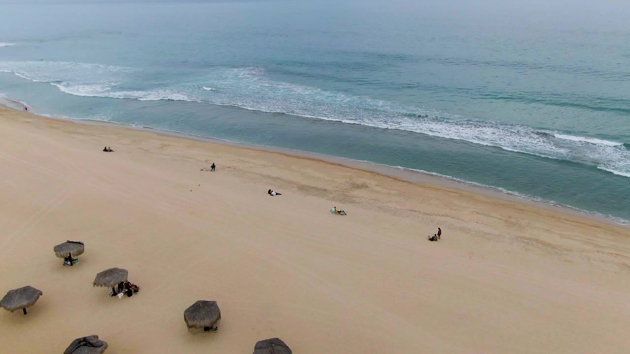 Aerial view over a San Jose del Cabo sandy beach with thatched umbrellas at the lower left, small people on the sand, and blue-green surf beyond.
