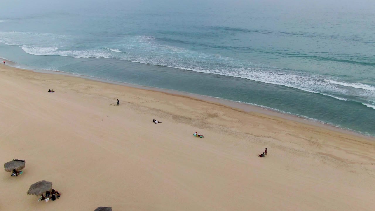 Aerial beach view with waves, scattered people, and umbrellas near the lower edge.