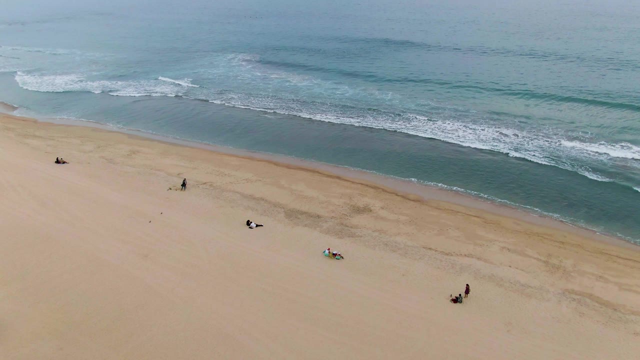 Aerial view of a San Jose del Cabo shoreline where the umbrellas are mostly out of frame, small people remain on the sand, and white surf advances toward shore.