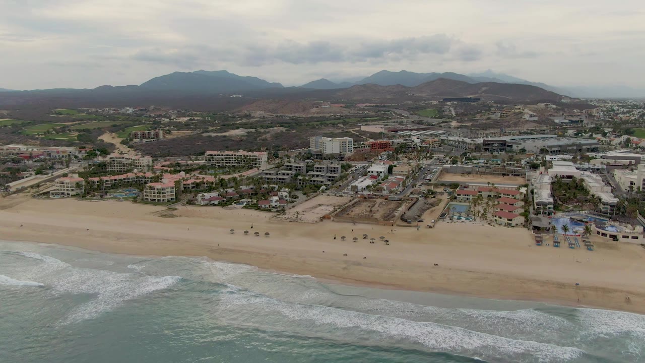 Aerial opening frame over San Jose del Cabo showing foamy surf in the foreground, a wide sandy beach, coastal buildings, distant mountains, and no visible people.