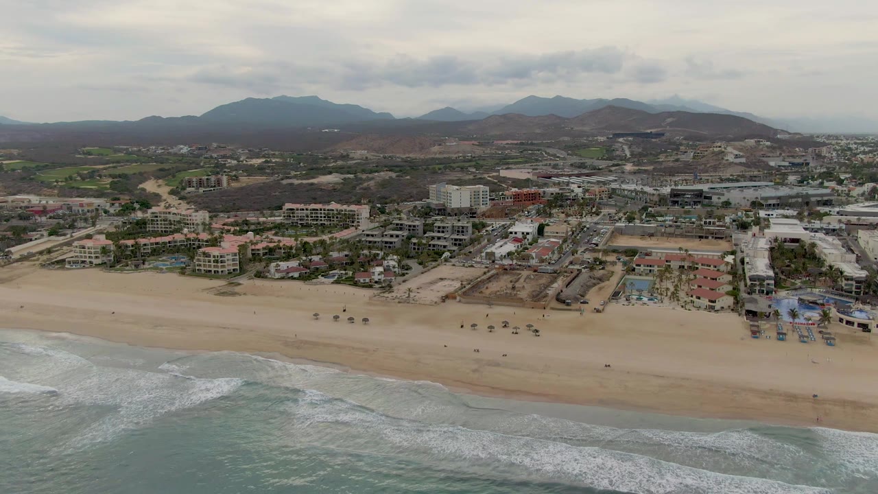 Second frame of the San Jose del Cabo coastline with wave foam shifted along the lower edge, tan beach across the middle, resort-area buildings behind it, and no visible people.