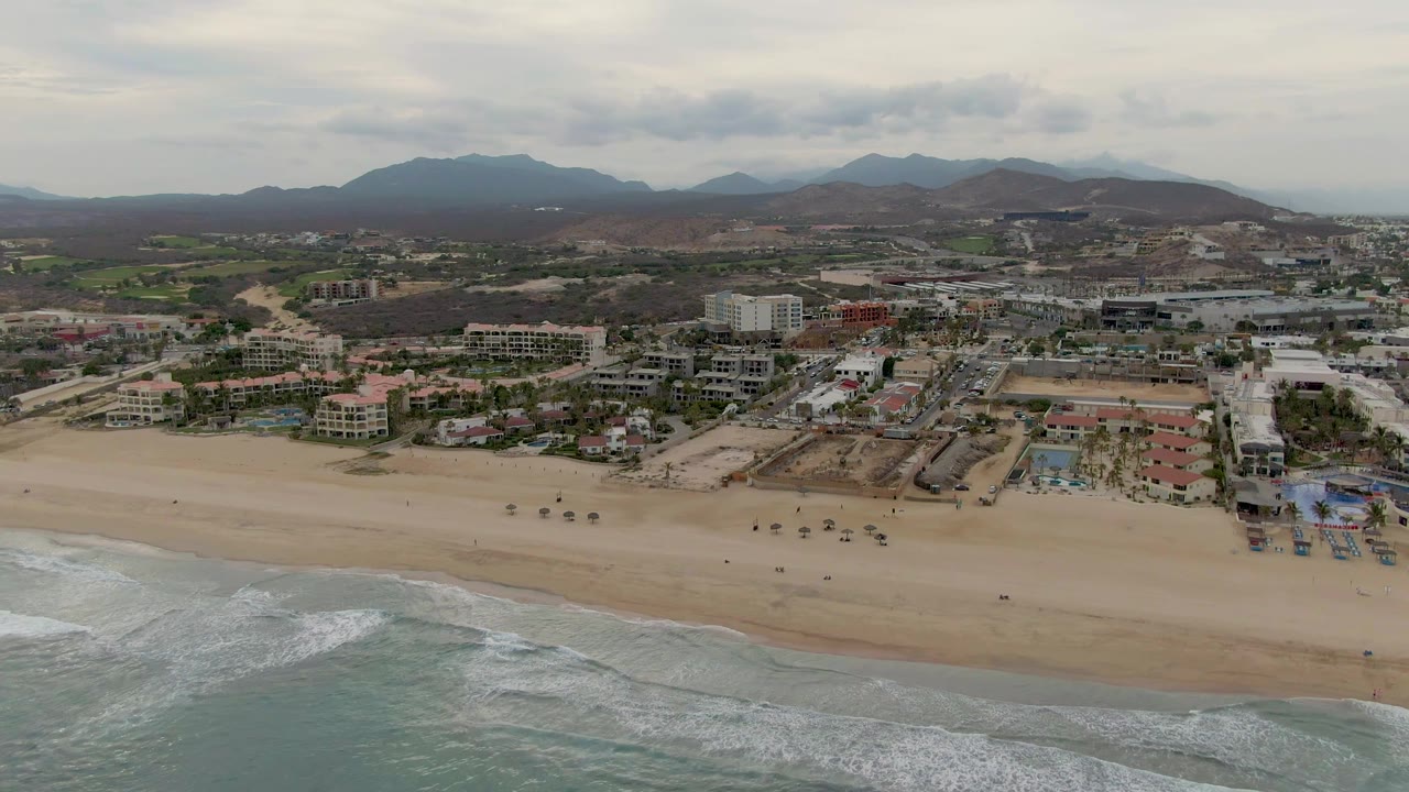 San Jose del Cabo aerial frame with rolling waves in the lower foreground, a mostly empty sandy beach, low coastal buildings, brown hills, distant mountains, and no visible people.