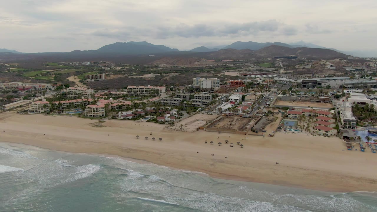 Aerial coastal panorama with surf, beach, shoreline buildings, and mountain horizon.