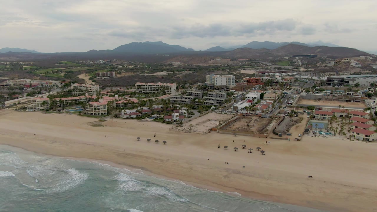 Opening aerial frame over San Jose del Cabo with ocean surf at the bottom, a wide sandy beach, resort buildings, roads, and distant mountains beneath an overcast sky.