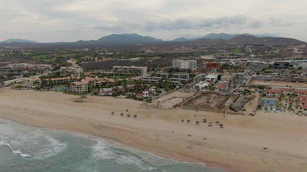 Wide beach shoreline with waves, umbrellas, buildings, and mountains.