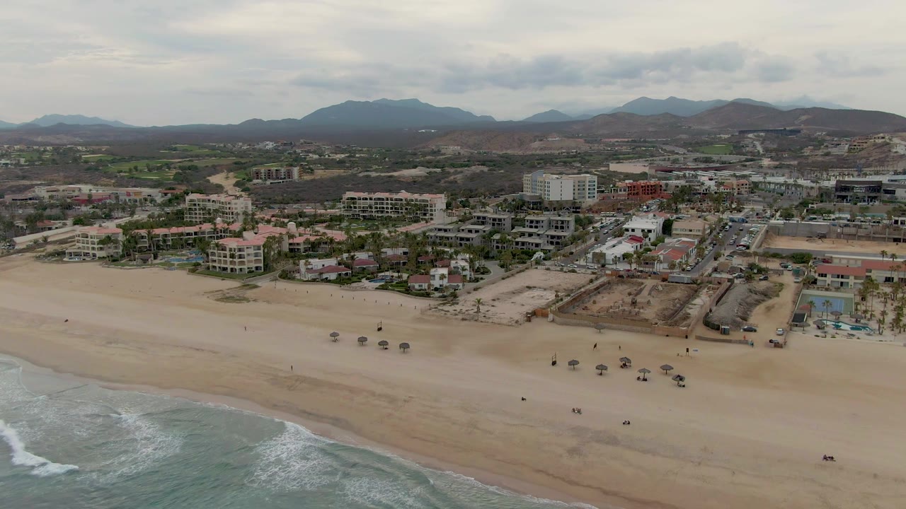 San Jose del Cabo aerial frame with a pale beach across the foreground, breaking waves at lower left, hotel-style buildings and roads in the middle, and hazy mountains across the horizon.