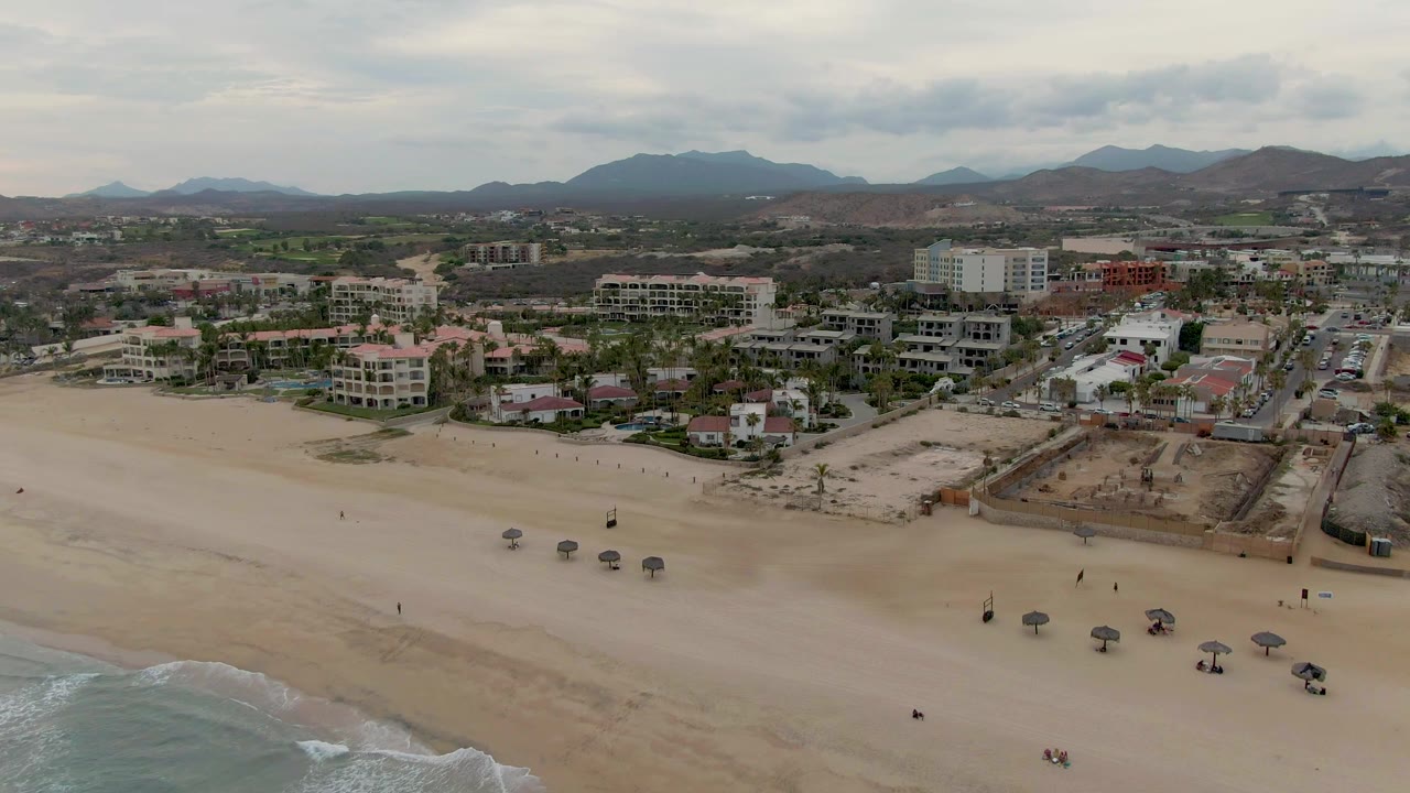 Aerial view of a sandy beach in San Jose del Cabo with ocean surf at lower left, several shade umbrellas on the sand, beachfront buildings, distant mountains, and small visible people.