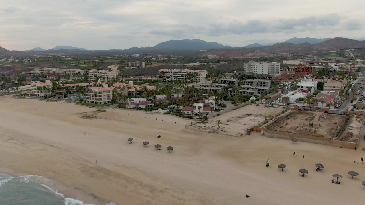 Wide drone view over beach sand with scattered umbrellas and coastal buildings.