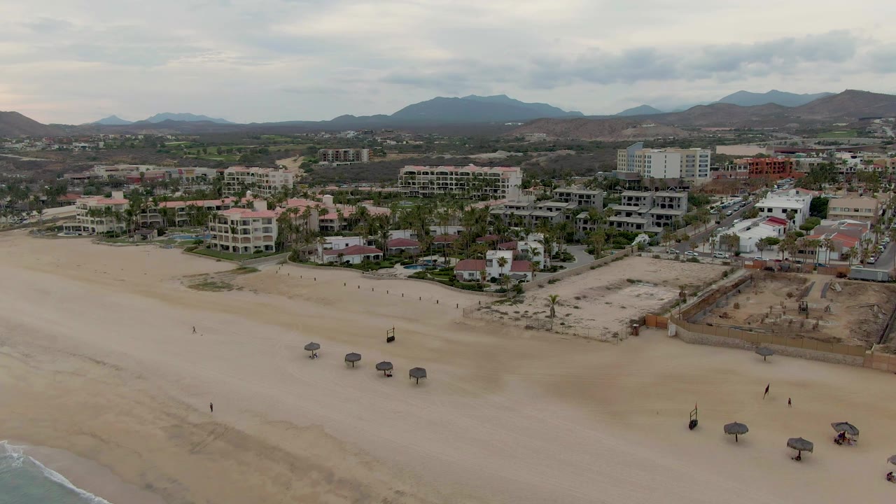 Aerial frame from San Jose del Cabo showing sparse beach visitors, a line of small shade umbrellas on the sand, low waves at the bottom left, and layered buildings with mountains beyond.