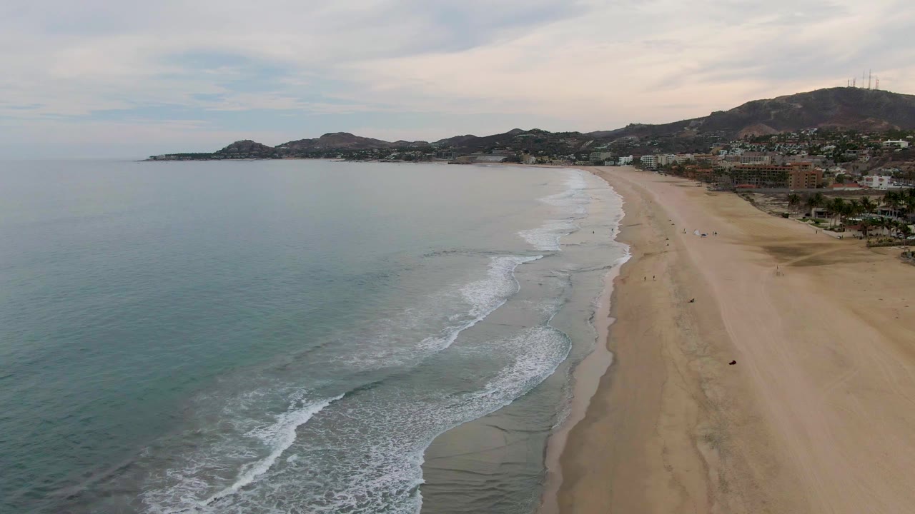 Aerial view over San Jose Del Cabo with blue-green ocean on the left, white surf along the sand, beachfront buildings to the right, distant hills, and small people visible on the beach.