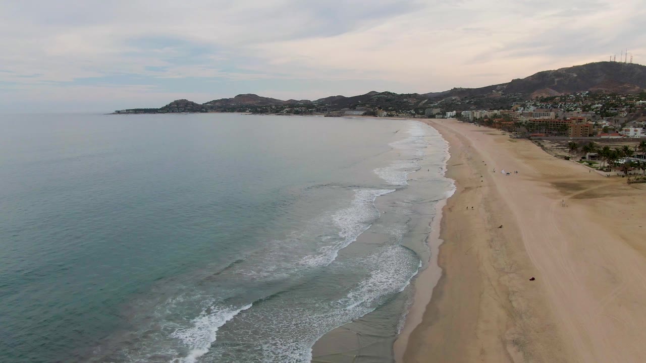 Drone view of blue-green water, breaking waves, sandy shoreline, buildings, and hills.