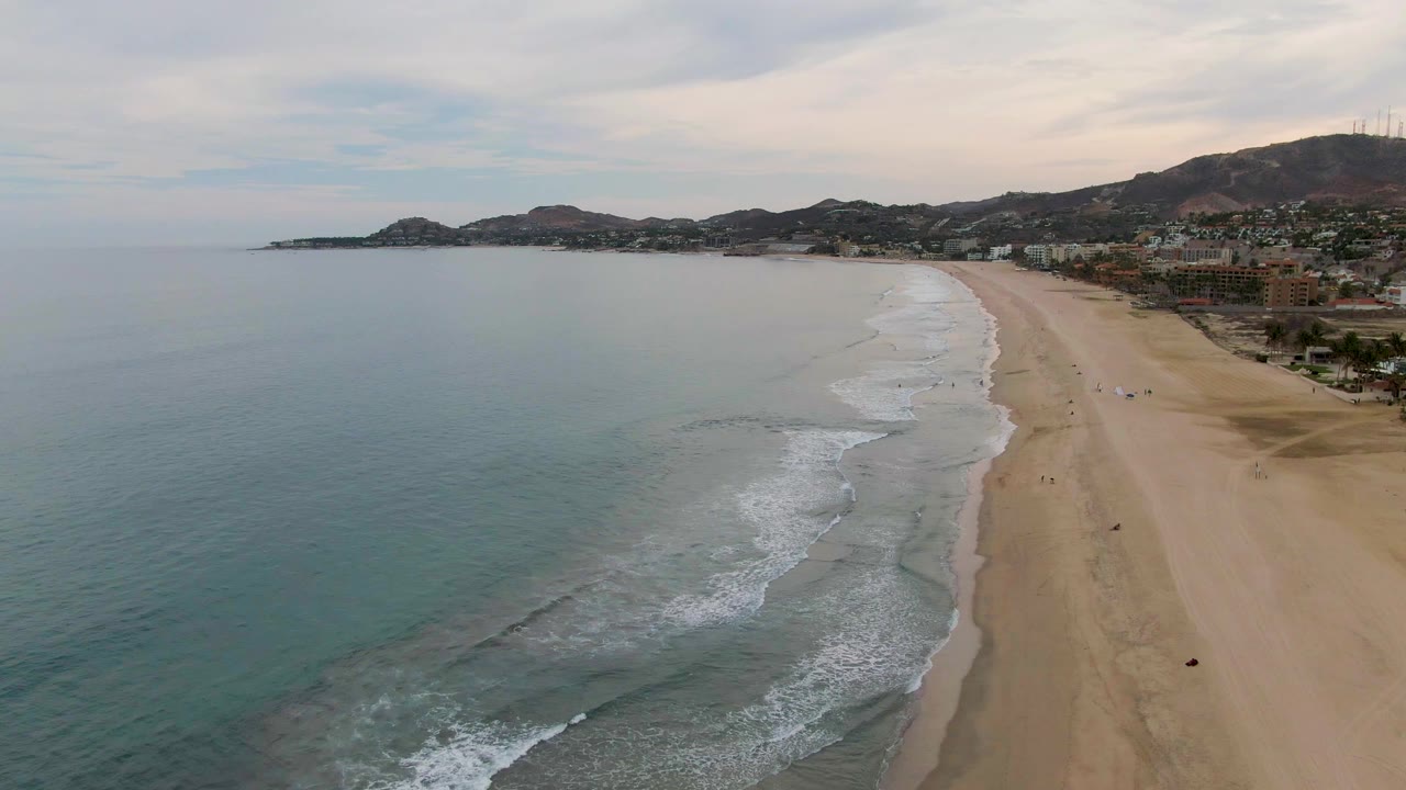 Wide aerial view of San Jose Del Cabo coastline with a broader curve of foam visible near the lower center, the sandy beach running up the right side, buildings beyond it, and tiny people on the sand.