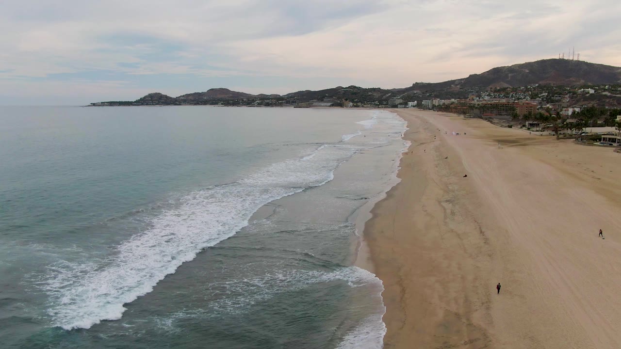 Aerial view over San Jose del Cabo beach, with blue-green ocean on the left, foamy surf entering the lower foreground, light sand on the right, distant hills, and tiny beachgoers visible.