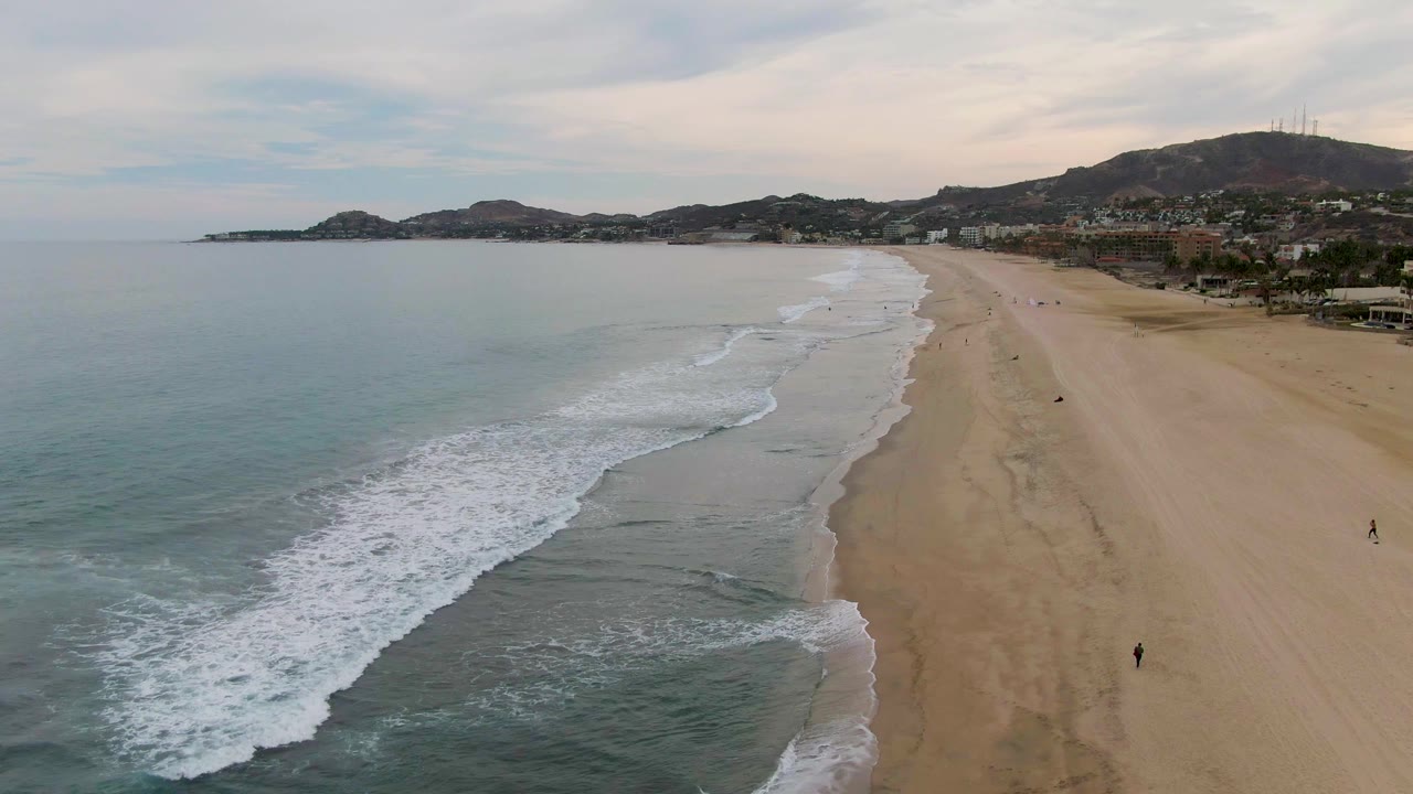 Foamy waves break along a wide sandy shoreline below distant hills.
