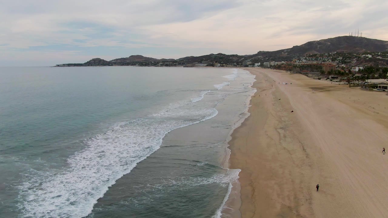 Aerial San Jose del Cabo beach view with the foam line pushed farther up the shore, calm blue water at left, pale sand at right, distant hills, and a few tiny people walking near the surf.