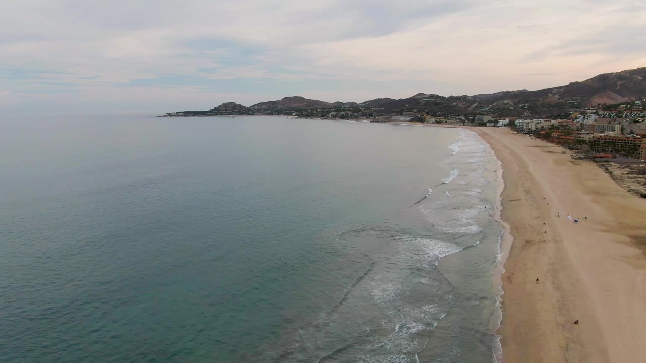 A high aerial frame over San Jose del Cabo shows blue-green ocean filling the left side, a sandy beach on the right, small shore waves, distant hills, coastal buildings, and tiny people along the shoreline.