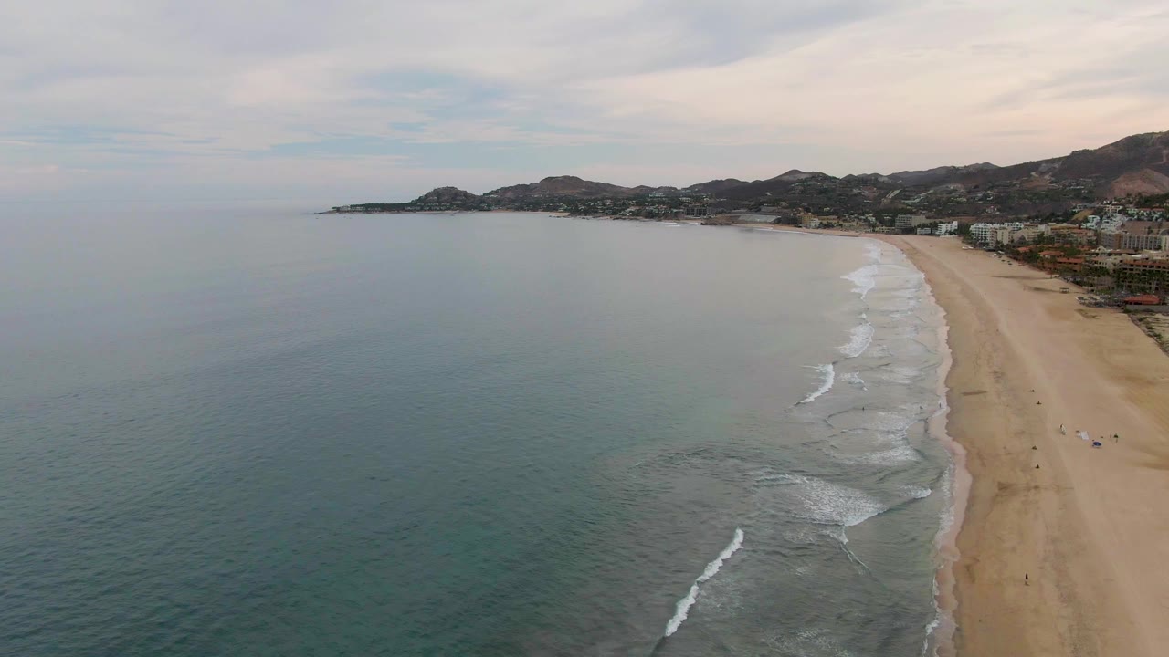 At 0:02, the ocean takes up most of the left frame while the sandy San Jose del Cabo beach narrows along the right edge, with white wave breaks, distant hills, buildings, and tiny figures visible.