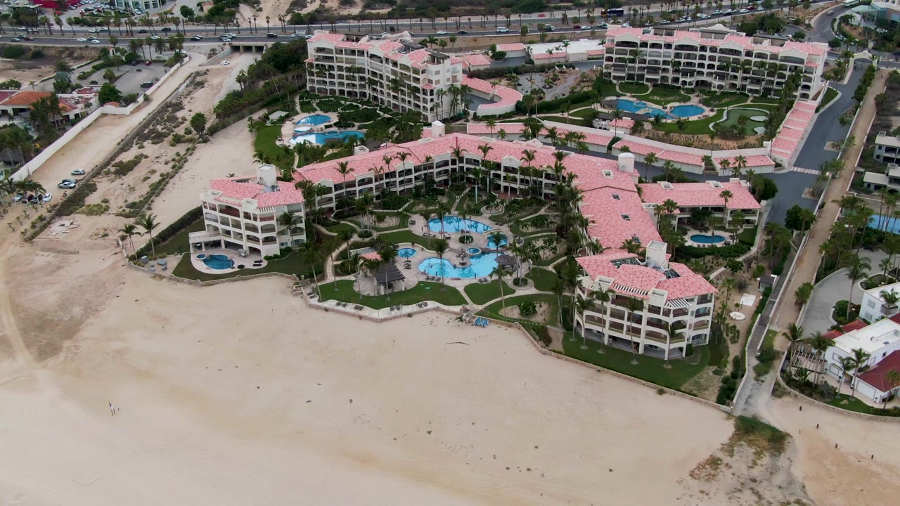 High aerial frame over a pink-roofed beachfront resort in San Jose Del Cabo, Mexico, with pools, landscaping, roads, hills, and no visible people.