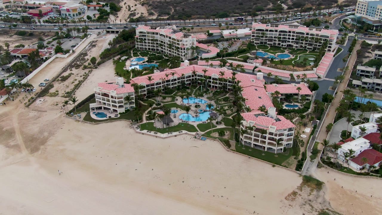 Aerial frame over the resort's central pools and pink roofs in San Jose Del Cabo, with the beach below and roads and hillside terrain behind, no visible people.