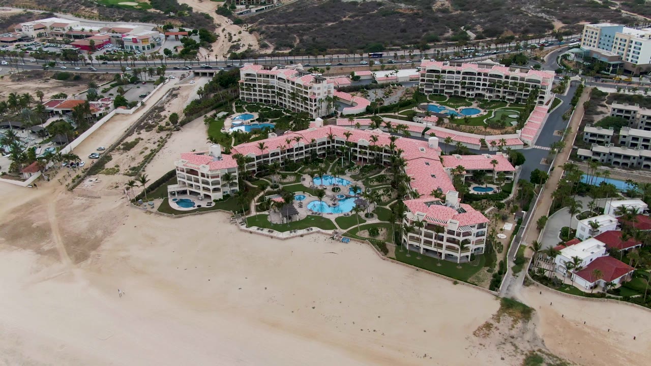 High drone frame of the San Jose Del Cabo resort complex, showing pale sand in the foreground, turquoise pools among pink-roofed buildings, and developed hills beyond, no visible people.