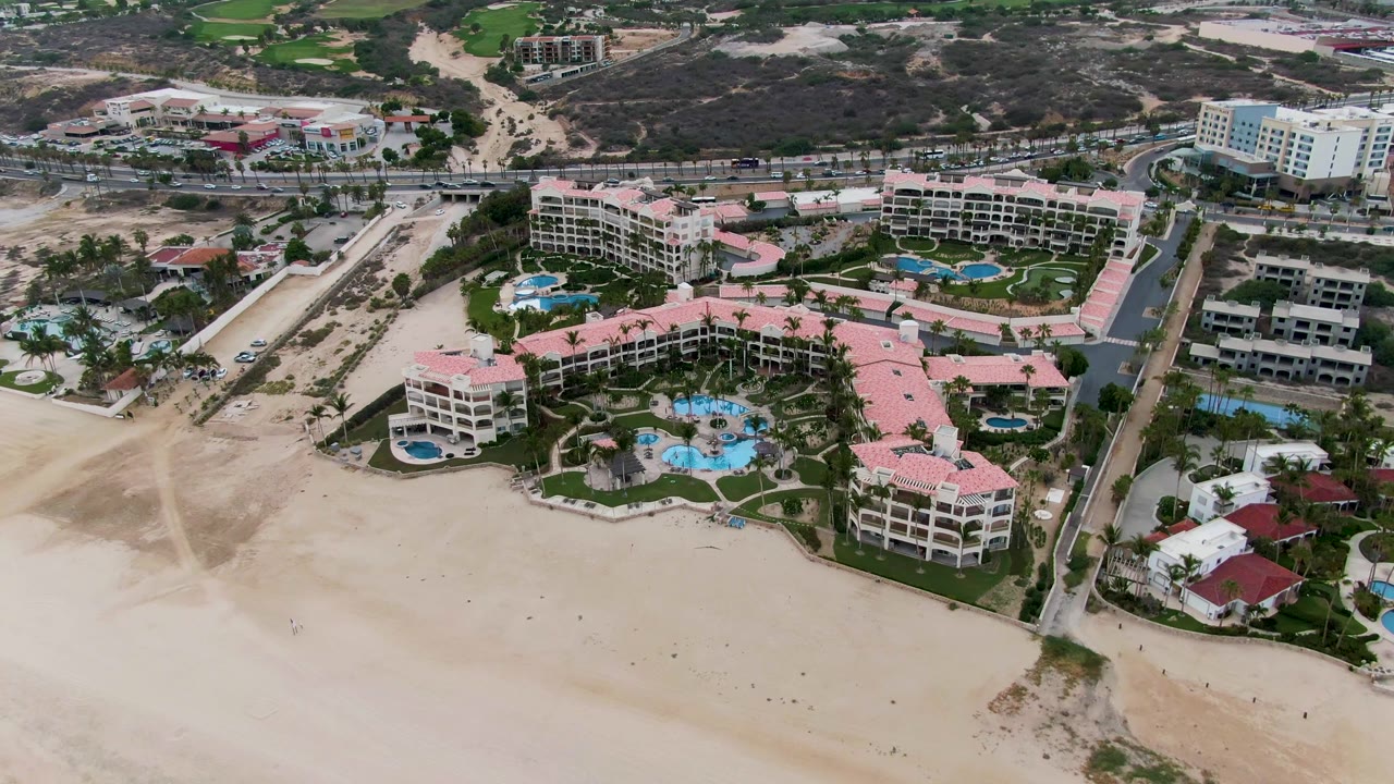 A later aerial frame in San Jose Del Cabo shows the resort from a slightly shifted angle, with more road and hillside development visible behind the pink roofs and pools, no visible people.