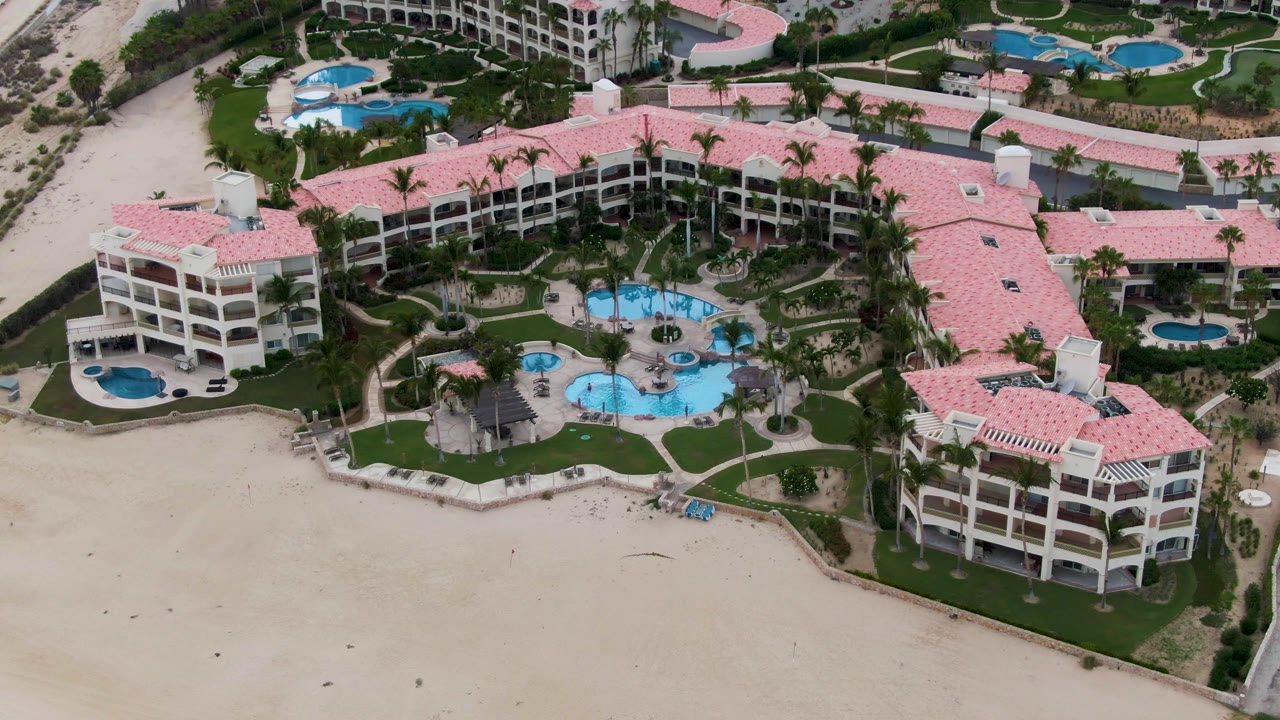 Aerial view of a San Jose del Cabo beachfront resort with pink tile roofs, blue pools, palm landscaping, sandy beach in front, and no visible people.