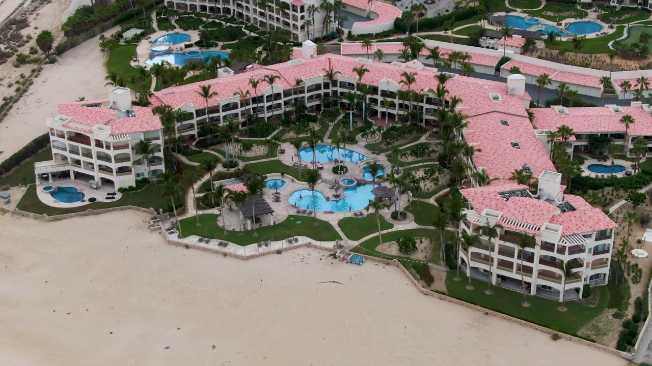 Elevated drone frame over the San Jose del Cabo resort, showing the central pool area, pink-roofed buildings, curved paths, beach sand, and no visible people.