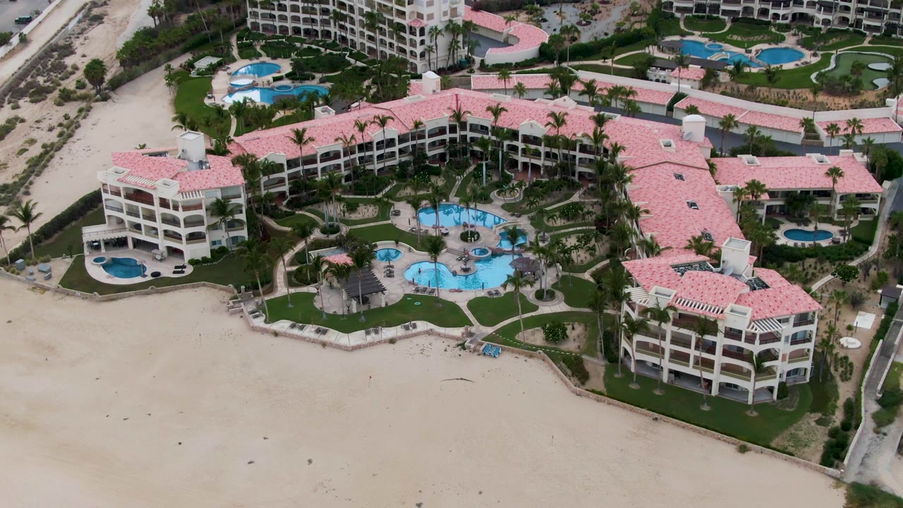 Aerial frame of the San Jose del Cabo resort with a wider look at adjacent buildings, blue pool shapes, palm-lined lawns, beach sand, and no visible people.