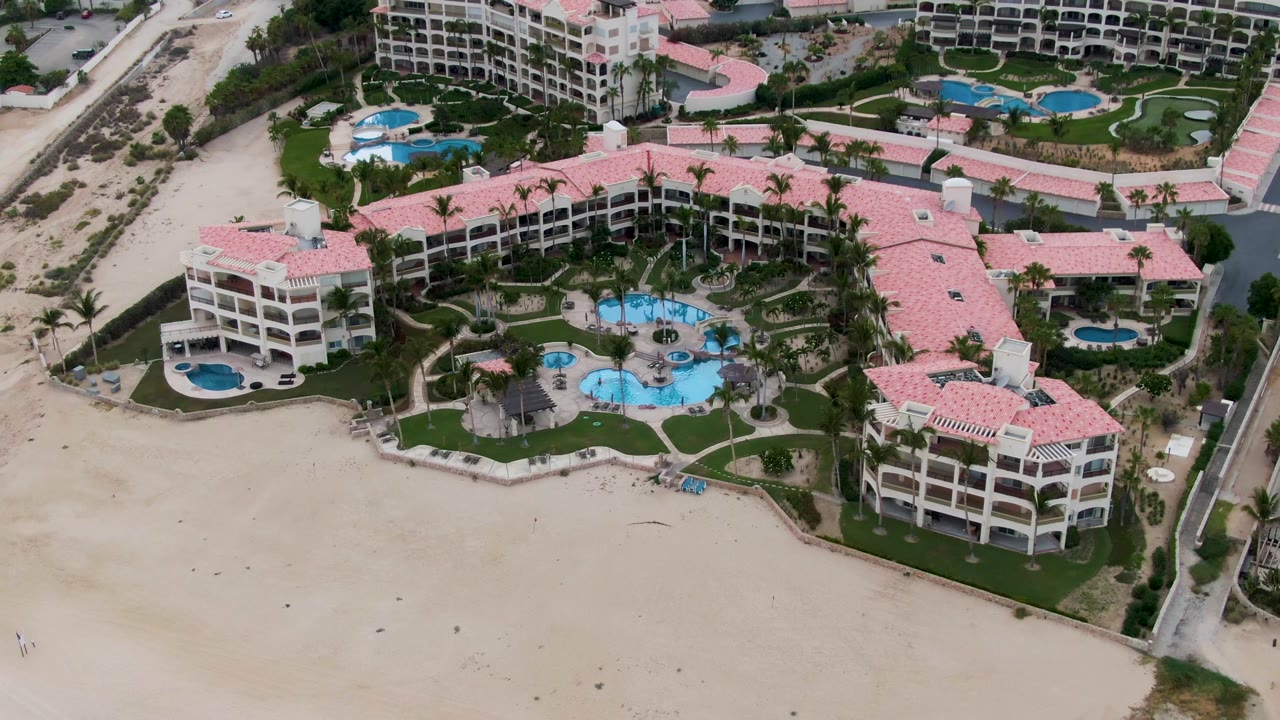 Drone view of beachfront resort, pools, lawns, and a road edge.