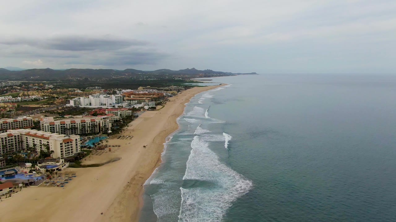 San Jose del Cabo coastline seen from above, with white wave lines moving toward the tan beach, multi-story coastal buildings on the left, muted ocean water on the right, and no visible people.