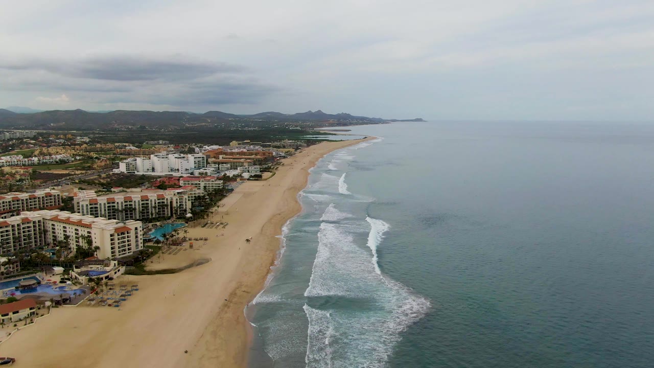 San Jose del Cabo aerial view with overcast light, beachfront buildings filling the lower left, pale sand running into the distance, surf curling near shore, calm ocean beyond, and no visible people.