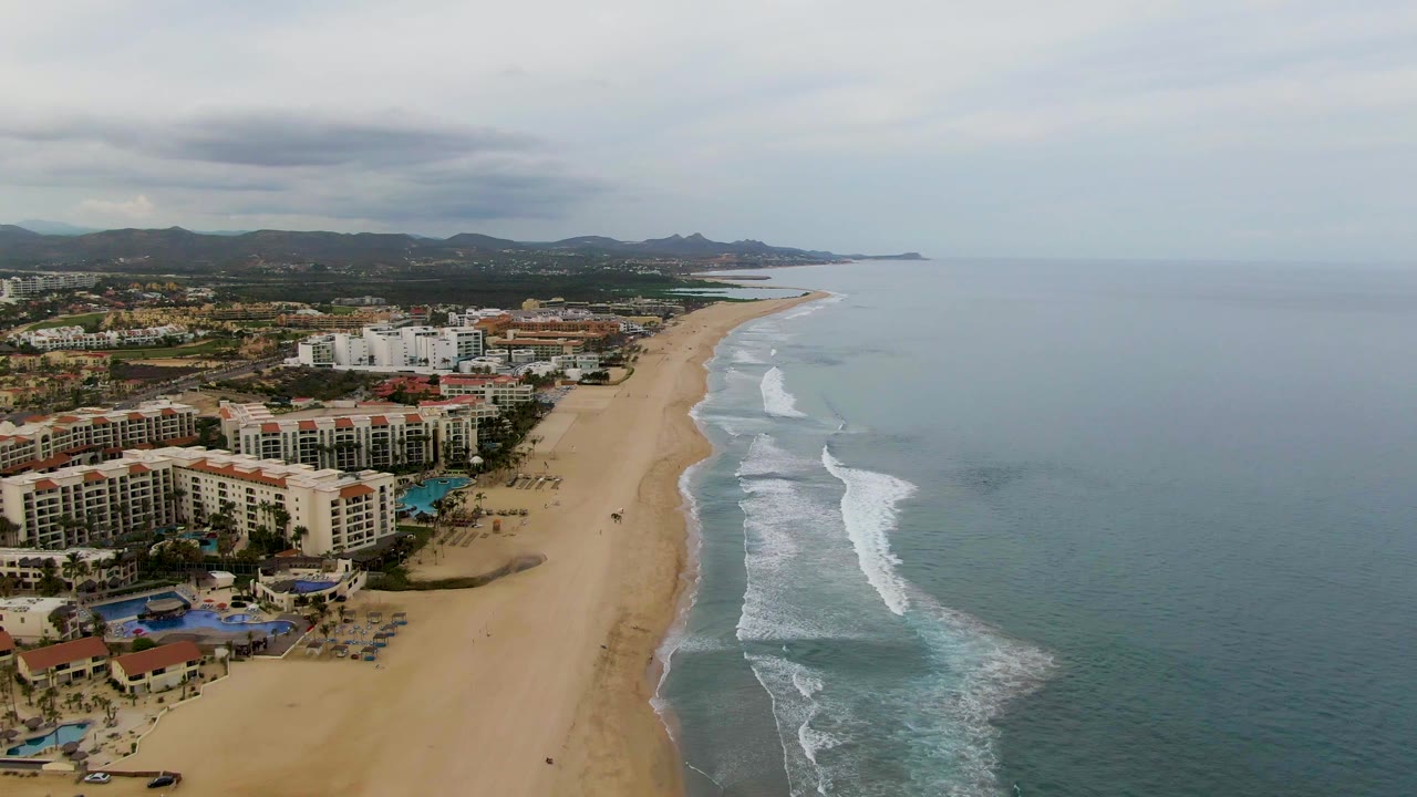Aerial view of resort hotels beside a sandy beach and breaking ocean waves.