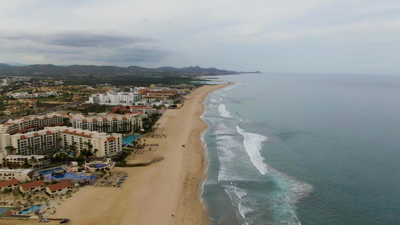 San Jose Del Cabo beachfront hotels and pools fill the lower frame while pale sand, white wave foam, blue-green water, and no visible people extend up the coast.