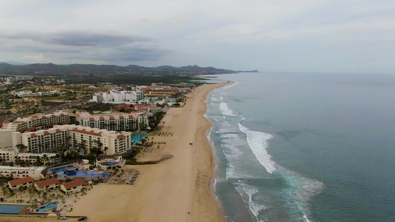 Wide aerial frame of the San Jose Del Cabo coastline with resort buildings at left, a long empty-looking beach, patterned surf bands, mountains, and no visible people.