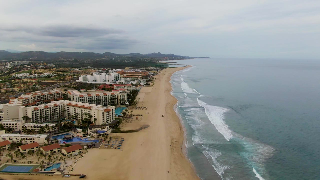 San Jose Del Cabo aerial view with resort blocks and pools closer in the foreground, surf breaking along the right side, hazy ocean horizon, and no visible people.