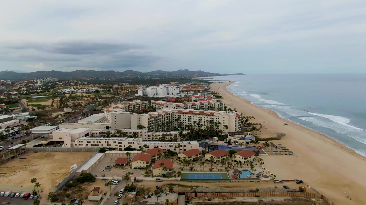 Aerial San Jose del Cabo frame showing cream hotel blocks, red-roofed buildings, turquoise pools, parked cars, surf lines, inland mountains, and no visible people.