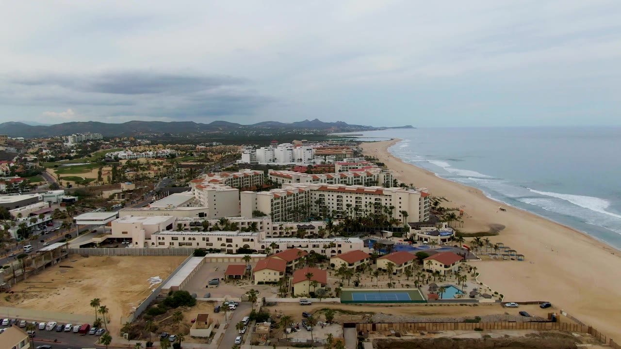 Wide aerial view in San Jose del Cabo with resort buildings in the lower frame, a broad sandy beach to the right, pale ocean surf, distant hills, and no visible people.