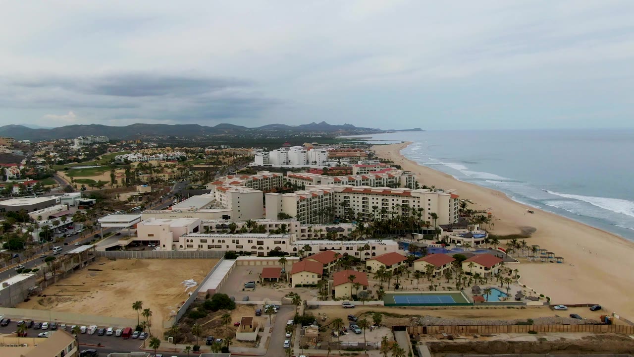 Mid-clip aerial view of San Jose del Cabo hotels near a road and parking area, with the sandy coast and ocean waves extending into the distance and no visible people.