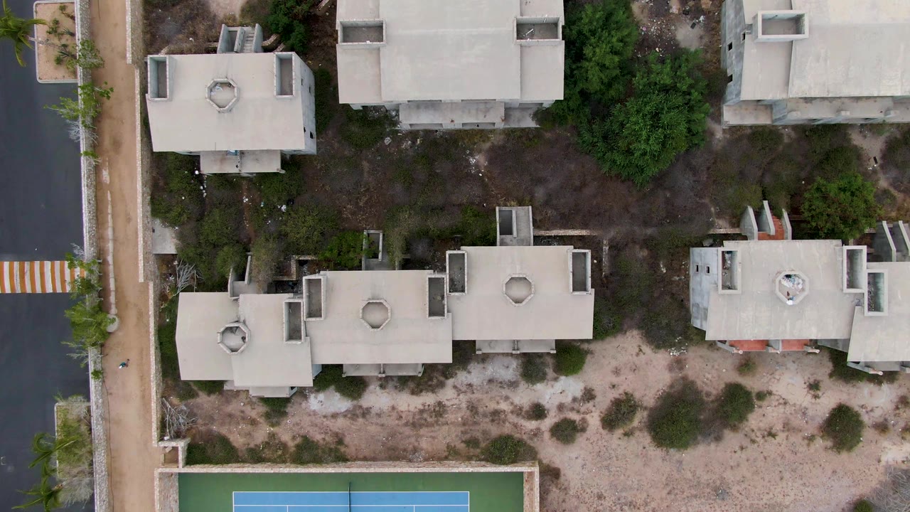 Second overhead frame in San Jose del Cabo with residential roofs centered, scrubby open ground below, road surfaces at both sides, and no visible people.