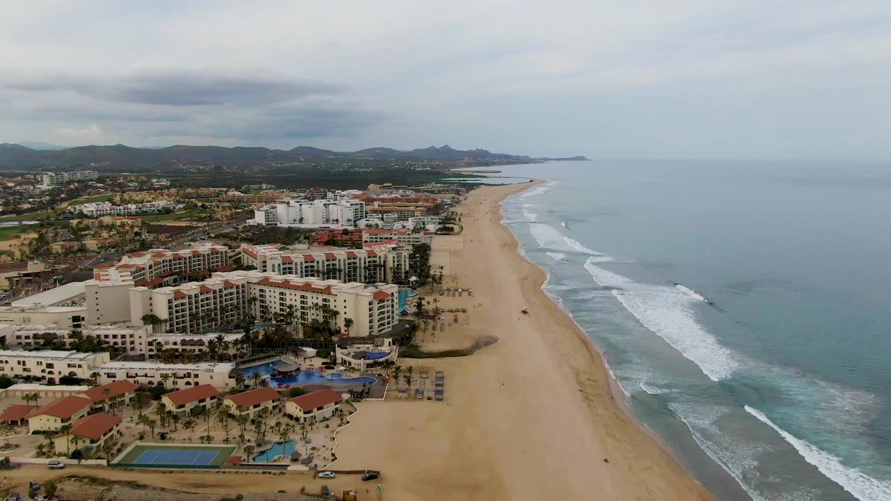 High aerial view in San Jose del Cabo showing resort buildings, a broad sandy beach, surf lines, distant mountains, cloudy sky, and no visible people.