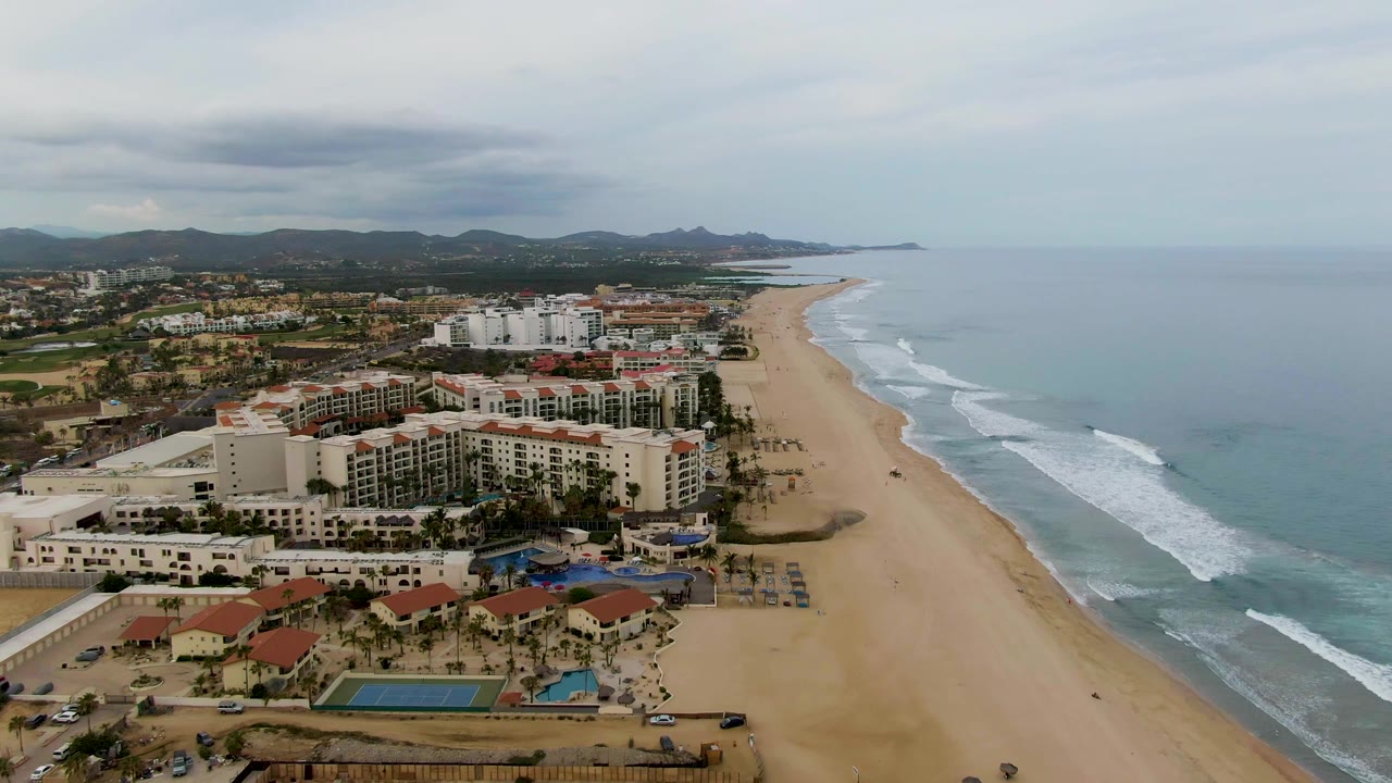 San Jose del Cabo coastline from above, with hotel buildings and palm grounds on the left, long sand beach through the center, ocean waves at right, and no visible people.