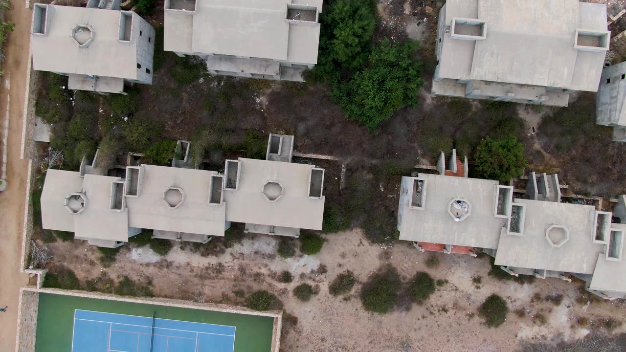Overhead frame in San Jose del Cabo showing flat concrete residential roofs, sandy ground, shrubs, and the upper edge of a blue tennis court, with no visible people.