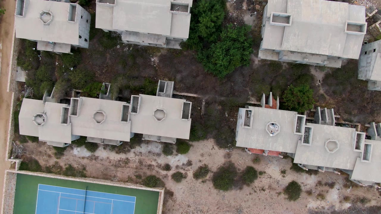 Aerial view of flat-roofed buildings beside scrub and a tennis court.