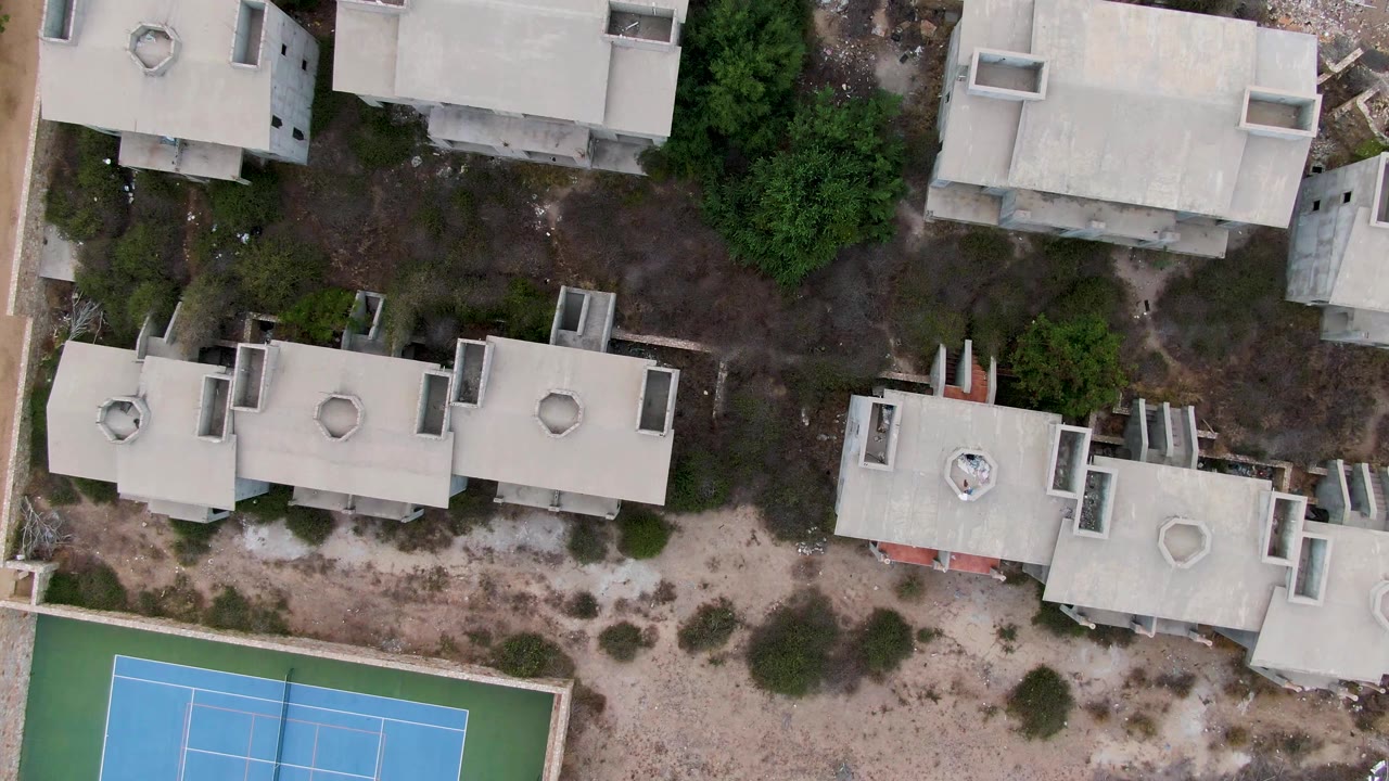 Top-down San Jose del Cabo frame with concrete roof shapes, sandy open ground, scattered bushes, trees near the upper right, and a blue tennis court at the bottom, with no visible people.