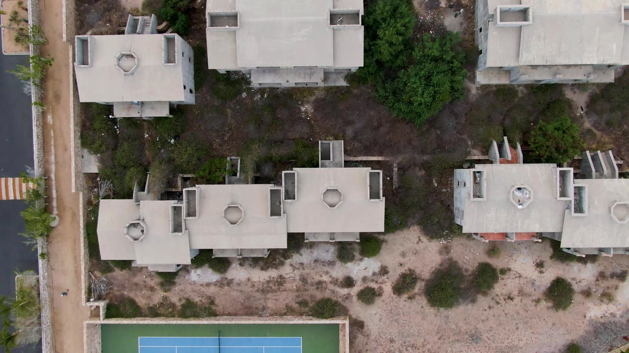 Top-down aerial frame over residential buildings in San Jose del Cabo, with pale roofs, sandy ground, shrubs, paths, and a partial blue and green tennis court at the bottom, no visible people.