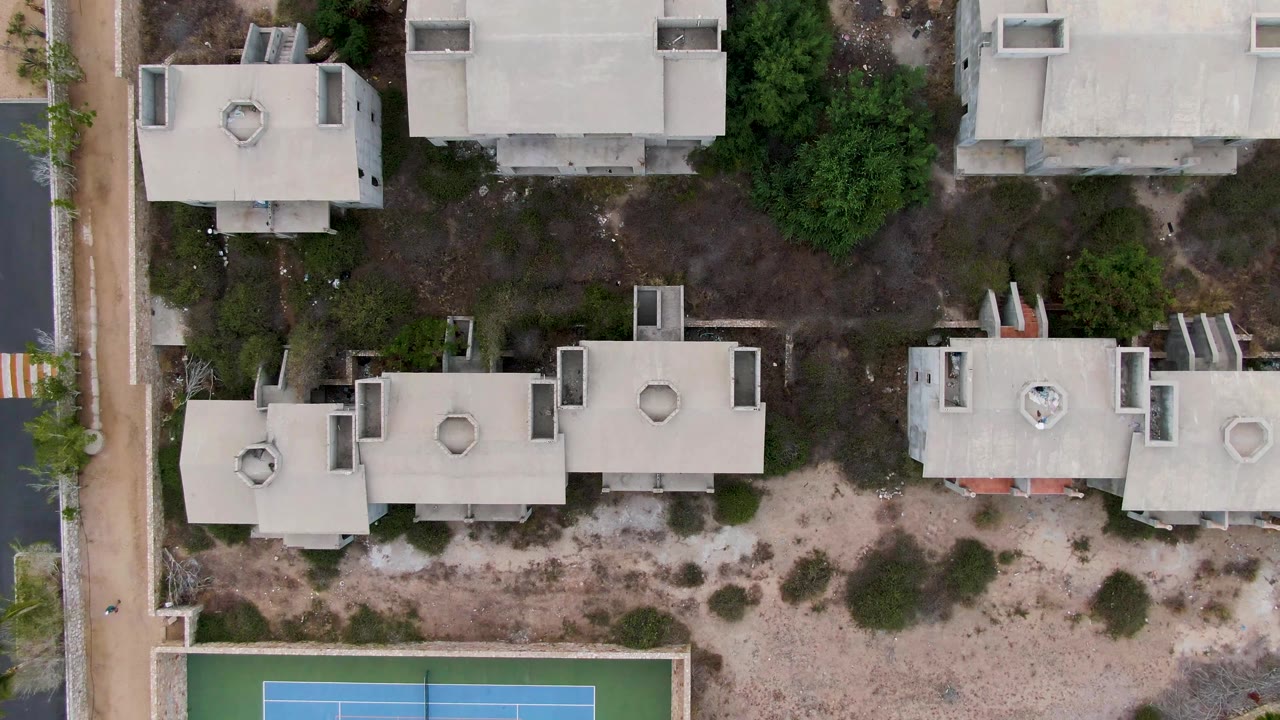 Second overhead frame in San Jose del Cabo showing apartment rooftops, a sandy open strip with low bushes, paved walkways, trees, and the tennis court edge near the lower border, no visible people.